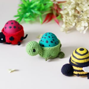 May include: Three decorative egg holders in the shape of insects. A red ladybug, a green turtle, and a yellow and black bee. Each holder cradles a painted egg, perfect for Easter or spring decor.