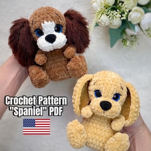 Könnte beinhalten: Zwei gehäkelte Spaniel-Hunde-Plüschtiere. Einer ist braun mit dunkelbraunen Ohren, der andere ist hellbraun. Beide haben blaue Augen und schwarze Nasen. Der Text "Crochet Pattern 'Spaniel' PDF" wird mit der amerikanischen Flagge angezeigt.