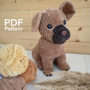 May include: A brown crocheted dog toy with black detailing around the eyes and nose. The toy is sitting on a white surface with a tan yarn ball and a brown yarn ball in the background. The text "PDF Pattern" is visible in the top left corner of the image.