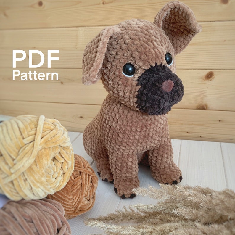 May include: A brown crocheted dog toy with black detailing around the eyes and nose. The toy is sitting on a white surface with a tan yarn ball and a brown yarn ball in the background. The text "PDF Pattern" is visible in the top left corner of the image.
