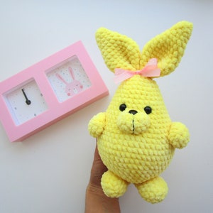 Op de afbeelding: Een gele gehaakte bunny met een roze strik. De bunny heeft zwarte ogen en een zwarte neus. De bunny zit op een wit oppervlak.