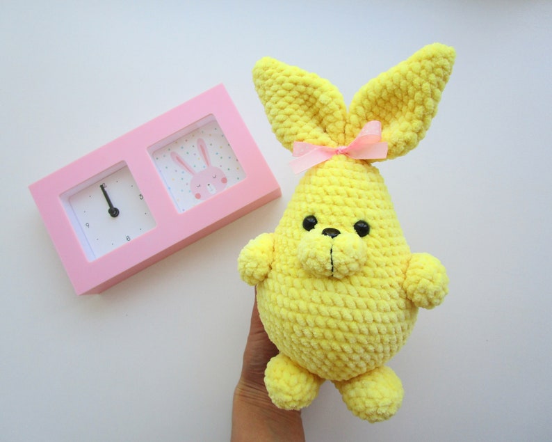 Op de afbeelding: Een gele gehaakte bunny met een roze strik. De bunny heeft zwarte ogen en een zwarte neus. De bunny zit op een wit oppervlak.