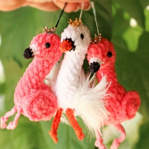 May include: Three crocheted stuffed toys: a white swan with a gold crown and two pink flamingos with gold crowns. The swan has white feathers and the flamingos have pink feathers.
