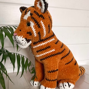 Puede incluir: Un peluche de tigre naranja con rayas negras y detalles blancos. Este juguete hecho a mano tiene un aspecto suave y texturizado, con hocico y patas blancas. El tigre est&aacute; sentado erguido, con la cabeza girada hacia un lado.