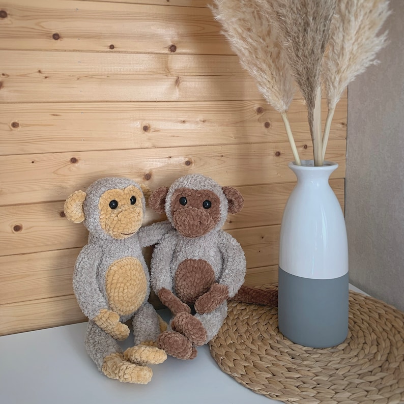 K&ouml;nnte beinhalten: Zwei geh&auml;kelte Affenspielzeuge mit braunen Augen und einem hellbraunen und beigefarbenen Farbschema. Die Affen sitzen auf einer geflochtenen Strohh&uuml;tte vor einer Holzwand. Eine wei&szlig;e Vase mit grauer Basis h&auml;lt getrocknetes Pampasgras.