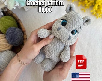 May include: A crocheted gray hippo plushie with blue eyes is held in two hands. The text "Crochet pattern Hippo" is above the hippo. A PDF icon and a US flag are also visible.