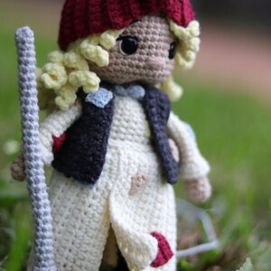 May include: A crocheted doll with blonde hair, wearing a white dress, a red bow, and a dark blue vest. The doll is holding a broom and is standing in a grassy area.