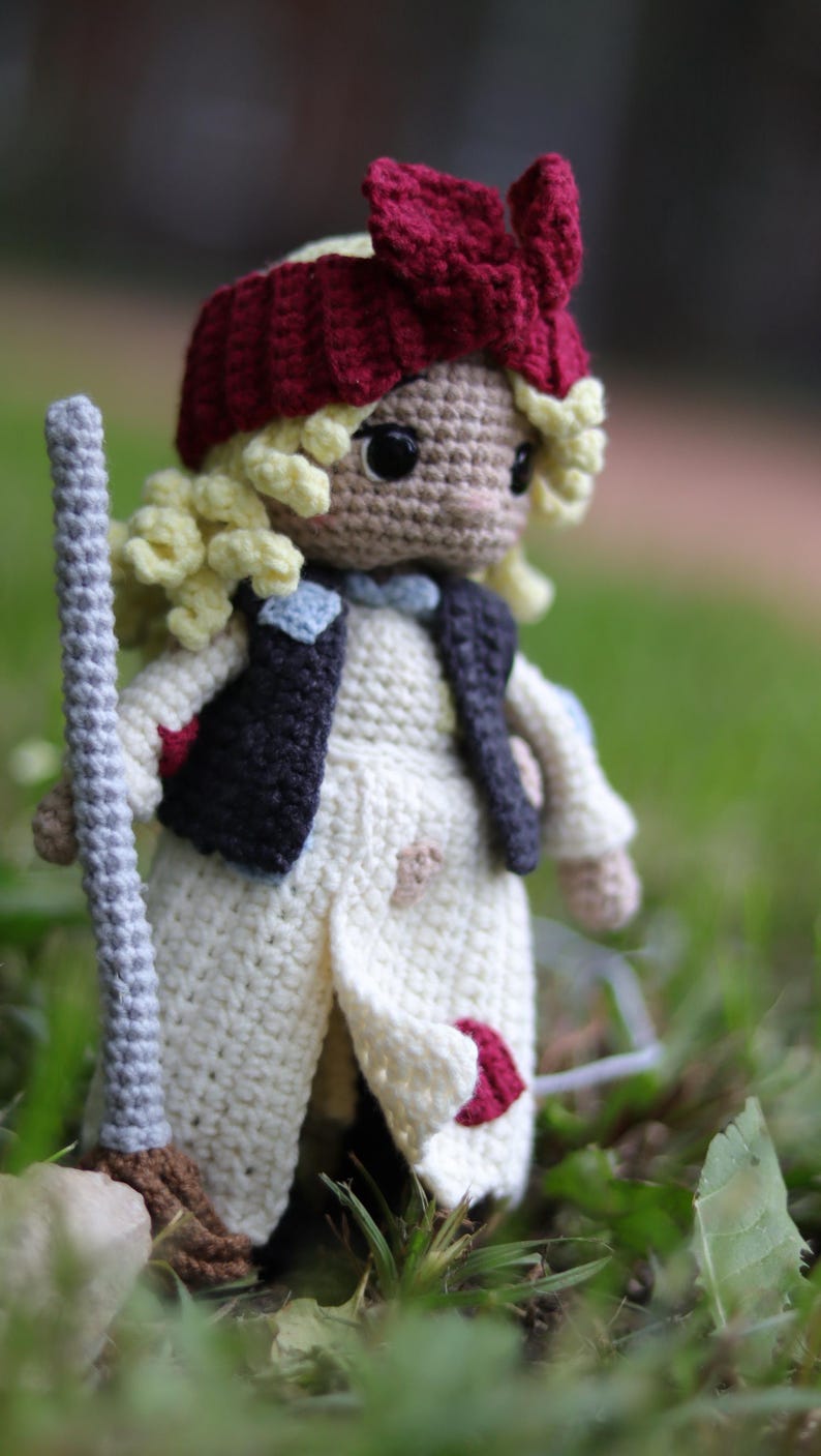 May include: A crocheted doll with blonde hair, wearing a white dress, a red bow, and a dark blue vest. The doll is holding a broom and is standing in a grassy area.