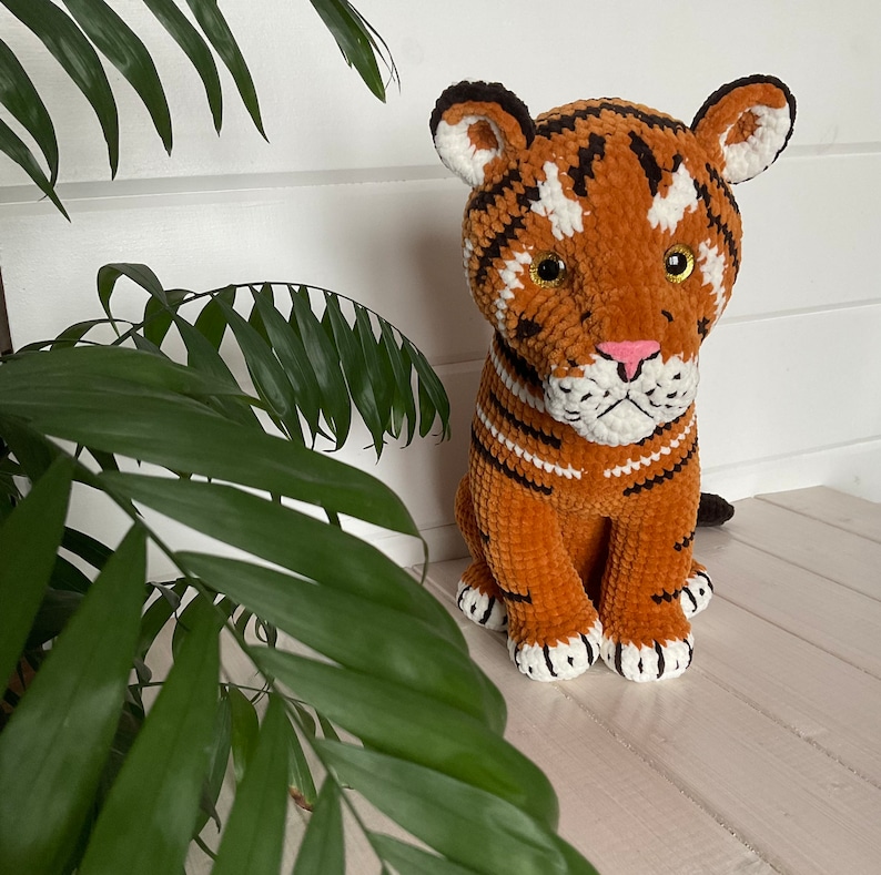 Puede incluir: Un peluche de cachorro de tigre hecho a mano. El juguete presenta rayas naranjas, negras y blancas, con una nariz rosa y ojos dorados. El tigre est&aacute; sentado y est&aacute; hecho de hilo suave.