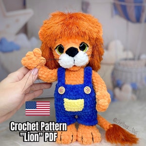 May include: Crocheted lion plush toy wearing blue overalls with yellow accents. The toy has a fluffy orange mane and tail. The image includes the text "Crochet Pattern "Lion" PDF" and a US flag.
