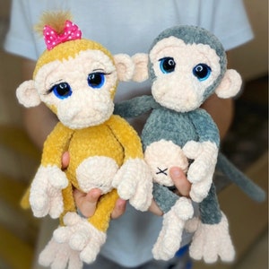 Può includere: Schema a uncinetto PDF per realizzare due giocattoli amigurumi a forma di scimmia. Una scimmia è gialla con un fiocco rosa e l'altra è grigia. Entrambe le scimmie hanno grandi occhi blu.