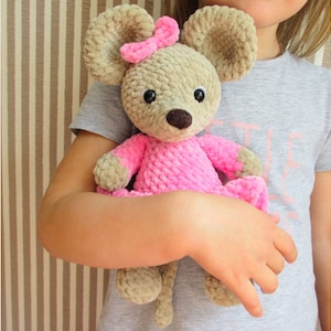 May include: A crocheted mouse toy wearing a pink dress and a pink bow. The mouse has brown eyes and a brown nose.