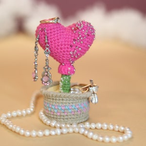 May include: A pink crochet heart-shaped jewellery holder with a green stem and a small basket base. The heart is decorated with silver beads and holds earrings and a ring. A pearl necklace is draped around the base.