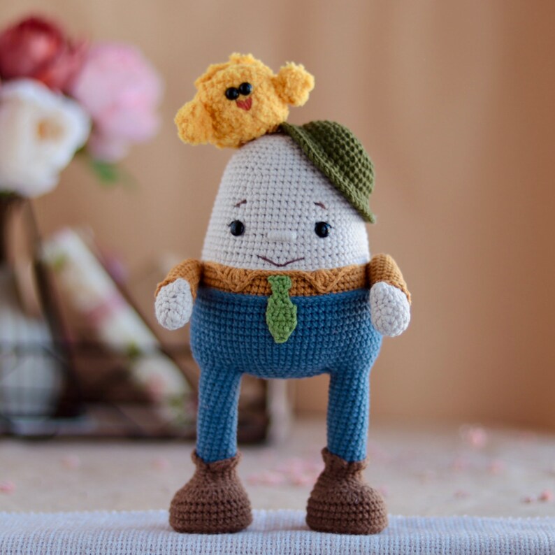 May include: A crocheted egg-shaped toy wearing a green hat and blue pants with a green tie. The toy has a friendly face and is holding a small yellow crocheted chick on its head.