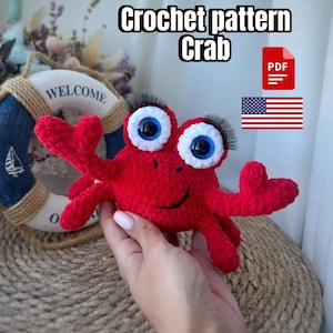 Pode incluir: Padrão de crochê para um brinquedo amigurumi de caranguejo vermelho com olhos brancos e um rosto sorridente. O padrão está disponível para download em PDF. A imagem mostra o brinquedo acabado sendo segurado em uma mão.