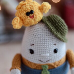 May include: A crocheted plush toy with a white body, brown arms, blue pants, and a green tie. The toy is wearing a green hat and has a yellow crocheted bird on its head.