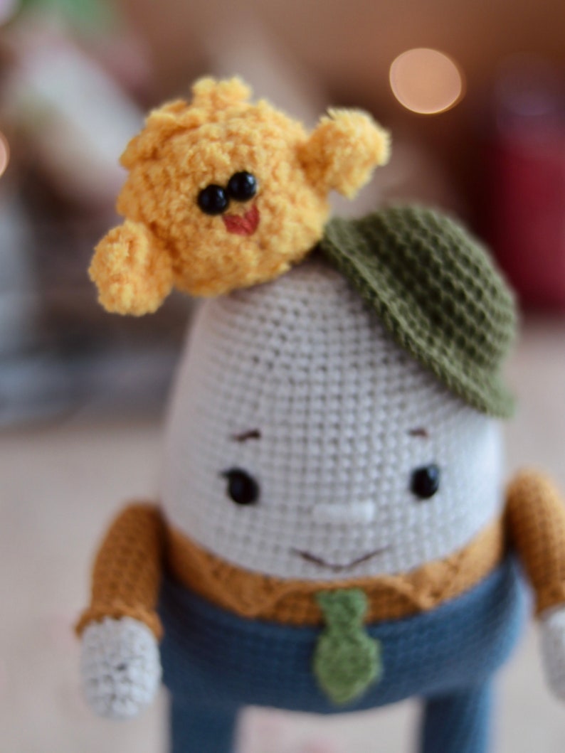 May include: A crocheted plush toy with a white body, brown arms, blue pants, and a green tie. The toy is wearing a green hat and has a yellow crocheted bird on its head.