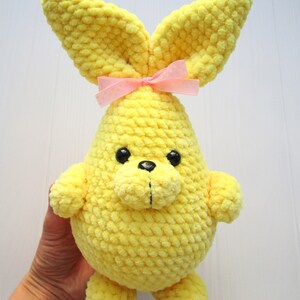 Op de afbeelding: Een gele gehaakte bunny met een roze strik. De bunny heeft een rond lichaam en lange oren.