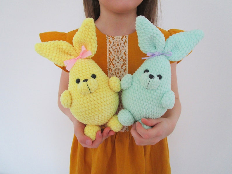 Op de afbeelding: Twee gehaakte konijnenknuffels, een gele en een lichtblauwe, vastgehouden door een kind. De konijnen hebben kleine zwarte ogen en roze en paarse strikjes.