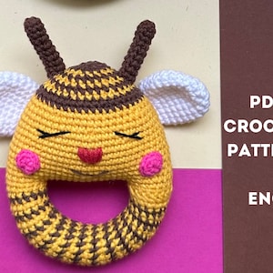May include: Crochet pattern for a bee-shaped baby rattle. The rattle is yellow and brown with pink cheeks and white wings. The pattern is in English and is available as a PDF download.