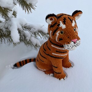 Puede incluir: Un juguete de tigre tejido a ganchillo con rayas naranjas y negras, sentado en la nieve. El tigre tiene una nariz rosa, detalles blancos y ojos dorados. Una rama de pino nevada est&aacute; en el fondo.