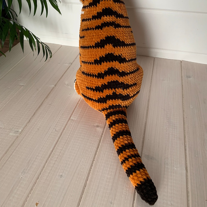 Puede incluir: Un juguete de tigre hecho a mano, de color naranja y negro. El cuerpo del tigre est&aacute; rayado con negro, y la cola es larga y rayada. El juguete est&aacute; hecho de hilo suave y texturizado. El tigre est&aacute; sobre una superficie de madera blanca.