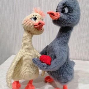 May include: Two crocheted stuffed animals, one yellow and one gray, holding a red heart. The animals are both birds with long necks and orange feet.