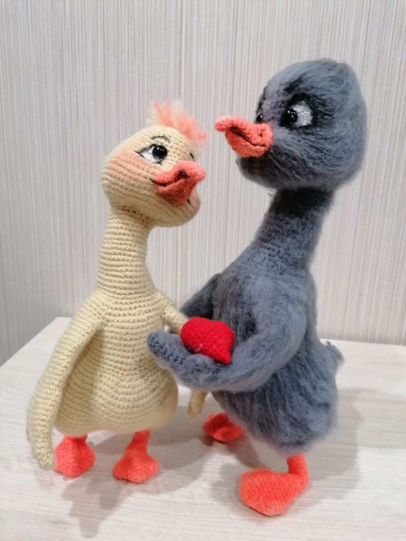 May include: Two crocheted stuffed animals, one yellow and one gray, holding a red heart. The animals are both birds with long necks and orange feet.