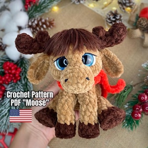 Puede incluir: Un juguete de alce de ganchillo con cuernos marrones, una bufanda roja y ojos azules grandes. El peluche es de color marrón y beige, con el texto "Crochet Pattern PDF 'Moose'" y una bandera americana.