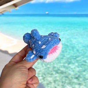May include: A handmade blue and white crochet whale shark with white spots and a pink mouth. The plush toy has black eyes and is held up against a backdrop of a turquoise ocean and blue sky.
