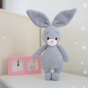 Puede incluir: Un juguete de conejo de crochet gris con nariz y ojos rosas. El conejo está de pie sobre una superficie blanca junto a un reloj rosa con una imagen de conejo.
