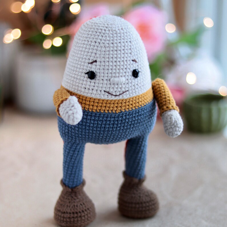 May include: A crocheted toy shaped like a person with a white head, yellow and blue clothing, and brown shoes.
