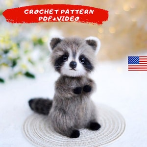 May include: Crochet pattern for a raccoon amigurumi toy. The pattern includes a PDF and video instructions. The raccoon is gray and black with a white face and is sitting on a white crocheted mat. The image also includes a small American flag.