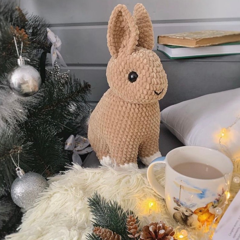 Op de afbeelding: Een gehaakt bruin konijn met witte pootjes zit op een wit, pluche oppervlak. Het konijn heeft zwarte ogen en een bruine neus. Er is een witte mok met een blauw-wit ontwerp en een kerstboom op de achtergrond.