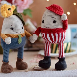 May include: Two crocheted egg-shaped toys with faces. One is wearing a red and white striped outfit and a red hat. The other is wearing a blue outfit with a brown hat and a yellow chick on top.
