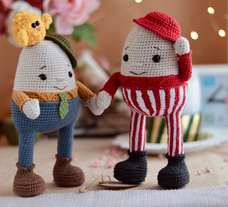 May include: Two crocheted egg-shaped toys with faces. One is wearing a red and white striped outfit and a red hat. The other is wearing a blue outfit with a brown hat and a yellow chick on top.