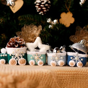 Könnte beinhalten: Ein Miniaturzug aus gehäkeltem Stoff mit verschiedenen farbigen Waggons. Jeder Waggon ist mit einem anderen Weihnachtsornament verziert, darunter eine Kiefernzapfen, ein Stern und eine Glocke. Der Zug steht auf einer beigefarbenen Strickdecke.