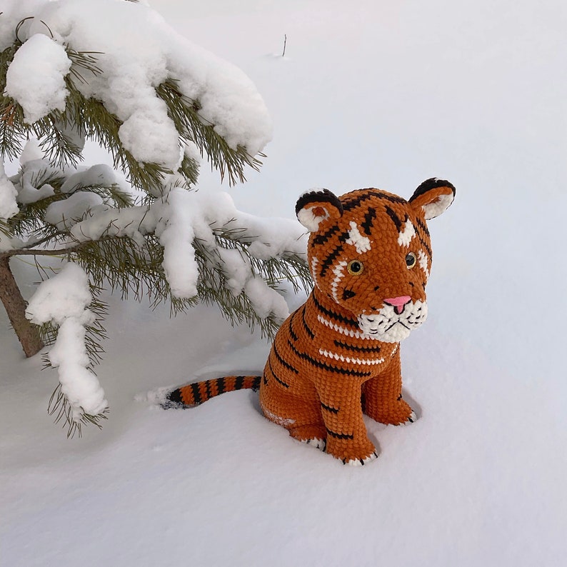 Puede incluir: Un cachorro de tigre de ganchillo con rayas naranjas y negras, sentado en la nieve cerca de una rama de pino nevada. El tigre tiene un hocico blanco, una nariz rosa y orejas con puntas negras. El fondo es un paisaje nevado.
