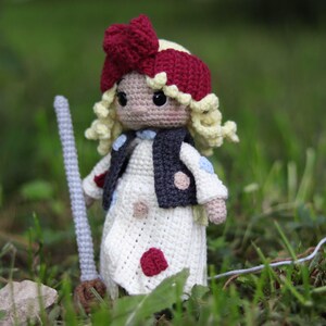 May include: A crocheted doll with blonde hair, a red bow, and a white dress with red and blue polka dots. The doll is holding a broom and is standing in a grassy area.