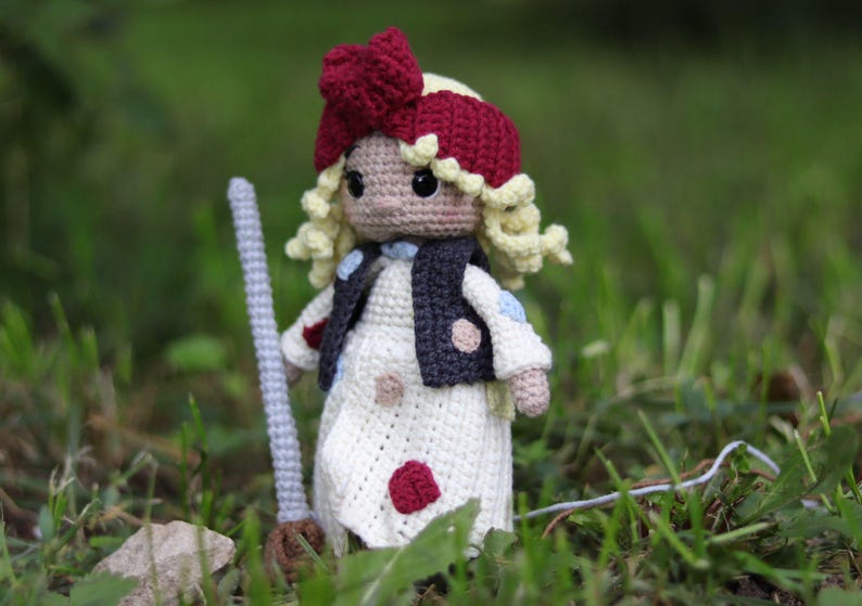 May include: A crocheted doll with blonde hair, a red bow, and a white dress with red and blue polka dots. The doll is holding a broom and is standing in a grassy area.