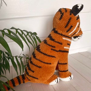 Puede incluir: Un juguete de tigre de peluche en posici&oacute;n sentada. El juguete es principalmente naranja con rayas negras y detalles blancos en las patas, la cara y la cola. El tigre tiene un aspecto suave y texturizado. Las orejas son marrones y blancas.