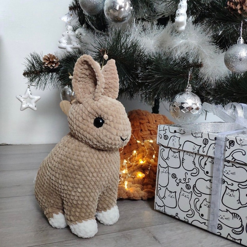 Op de afbeelding: Een gehaakt bruin konijn met witte voeten dat voor een kerstboom met ornamenten en een cadeaudoos met een kattenpatroon zit.