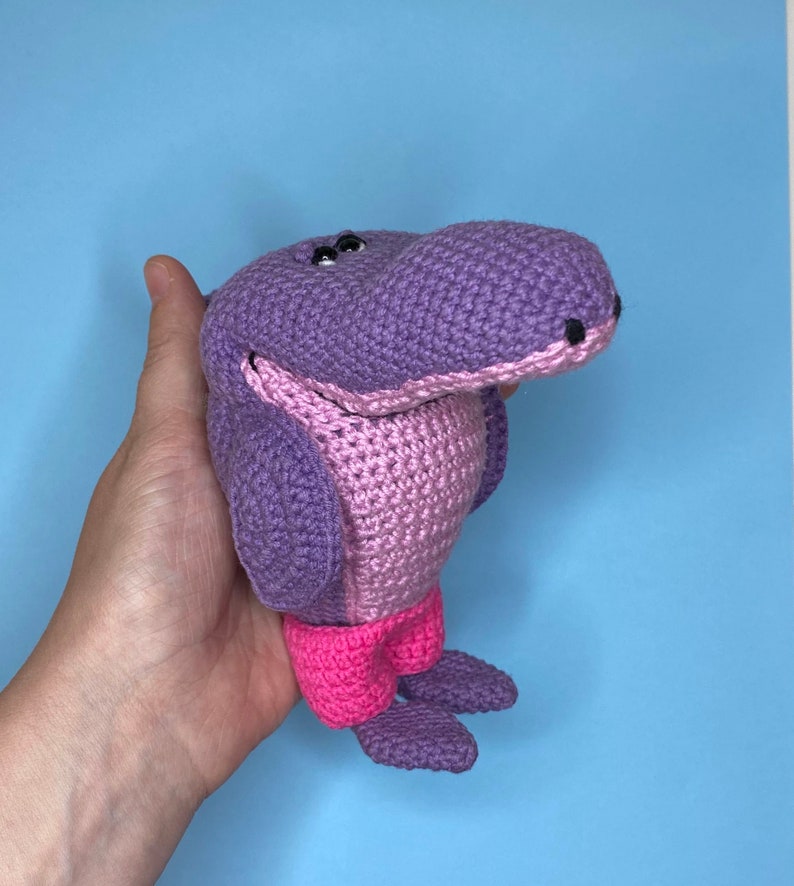 K&ouml;nnte beinhalten: Ein geh&auml;keltes lila und pinkfarbenes Nilpferd-Spielzeug mit schwarzen Augen. Das Nilpferd tr&auml;gt eine rosa Shorts.
