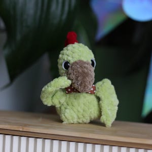 Puede incluir: Un pájaro de peluche verde claro hecho a mano con un pico marrón y una cresta roja. Tiene una pequeña pajarita de lunares rojos y blancos. El pájaro está sentado sobre una superficie de madera.