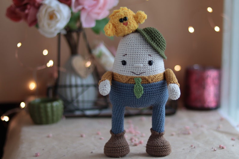 May include: A crocheted egg-shaped toy with a yellow chick on top, wearing a green hat and blue pants. The toy has brown shoes and a green tie.