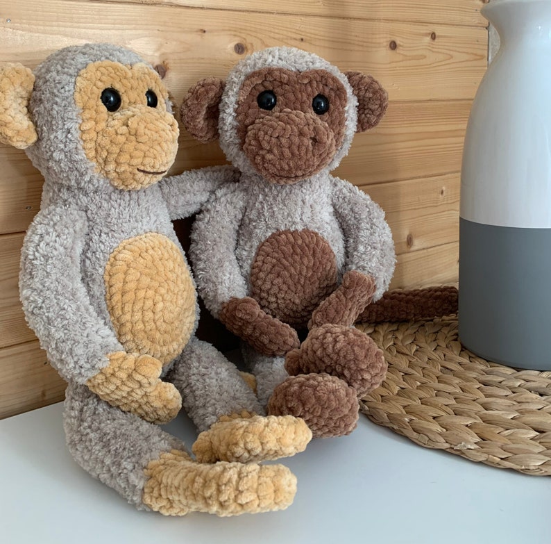 K&ouml;nnte beinhalten: Zwei geh&auml;kelte Affenspielzeuge, eines braun und eines beige, die auf einer geflochtenen Weidenoberfl&auml;che sitzen. Der braune Affe hat einen braunen Bauch und der beige Affe einen beigen Bauch.