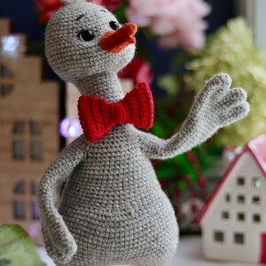 May include: A gray crocheted duck wearing a red bow tie. The duck has a small orange beak and orange feet. The duck is standing on a white surface.