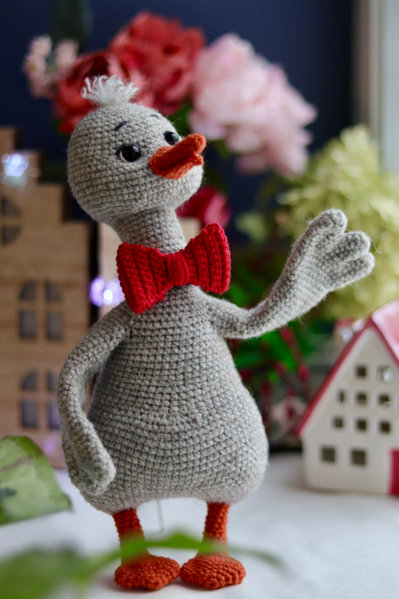 May include: A gray crocheted duck wearing a red bow tie. The duck has a small orange beak and orange feet. The duck is standing on a white surface.