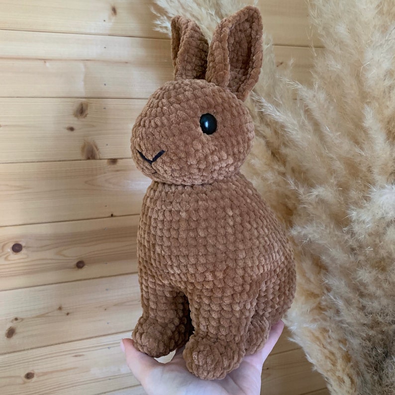 Op de afbeelding: Een bruin gehaakt pluche konijn met zwarte ogen. Het konijn zit op een hand.
