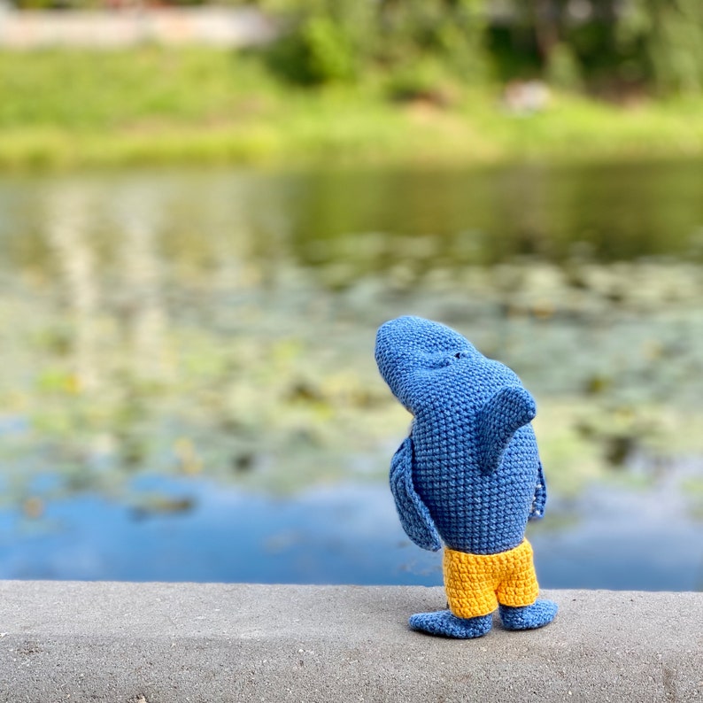 K&ouml;nnte beinhalten: Ein blaues, geh&auml;keltes Hai-Spielzeug mit gelben Shorts steht auf einer grauen Oberfl&auml;che und ist von der Kamera abgewandt. Der Hintergrund ist ein verschwommenes Bild eines Teichs oder Sees.